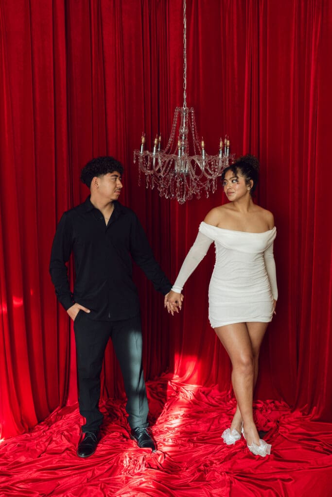 Couple holding hands beneath crystal chandelier and red velvet curtains during vintage Valentine’s Day mini session at LA Loft Studios in Los Angeles.