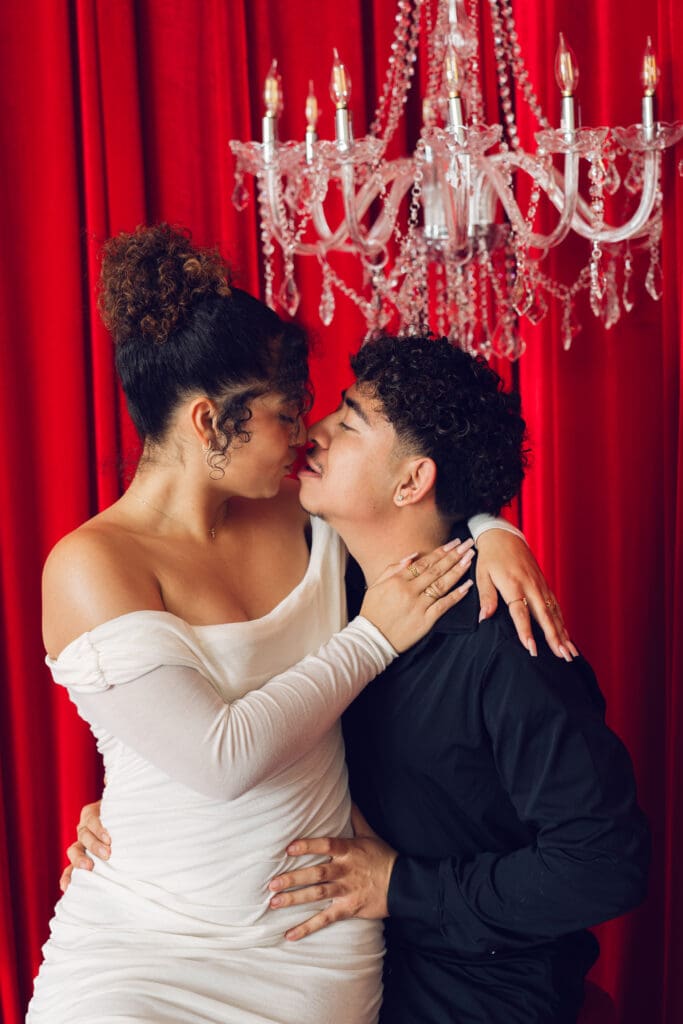 Close-up of romantic couple leaning in for a kiss beneath crystal chandelier and red velvet backdrop during Valentine’s Day photoshoot at LA Loft Studios in Los Angeles.
