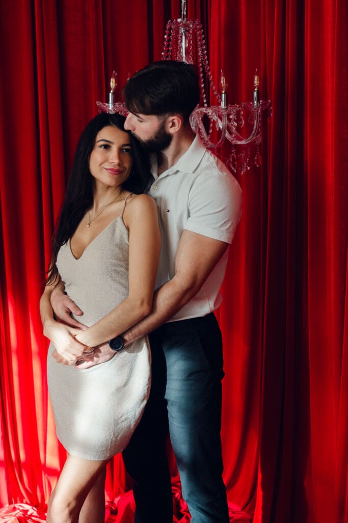 Couple embracing beneath crystal chandelier and red velvet curtains during romantic vintage Valentine’s Day mini session at LA Loft Studios in Los Angeles.